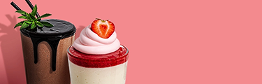 Two beverages are on a pink background. On the left is a chocolate shake with chocolate syrup dripping down the side and a sprig of mint on top. On the right is a layered dessert in a glass, with a white base, a red layer, pink whipped cream, and a strawberry on top.