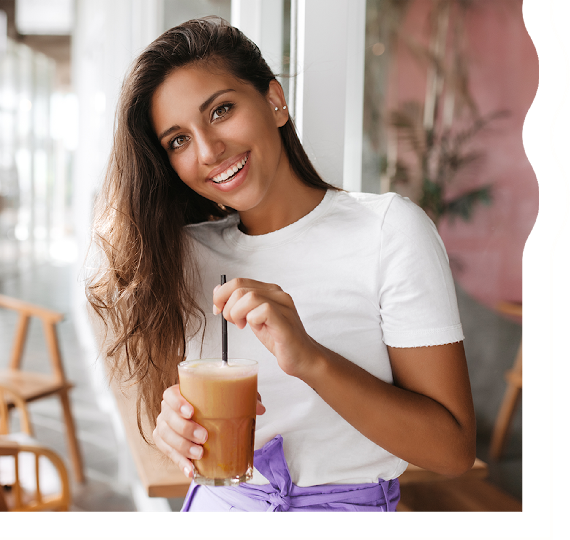 A woman with tan skin and long, dark brown hair is smiling and looking directly at the camera. She is holding a creamy chocolate shake.