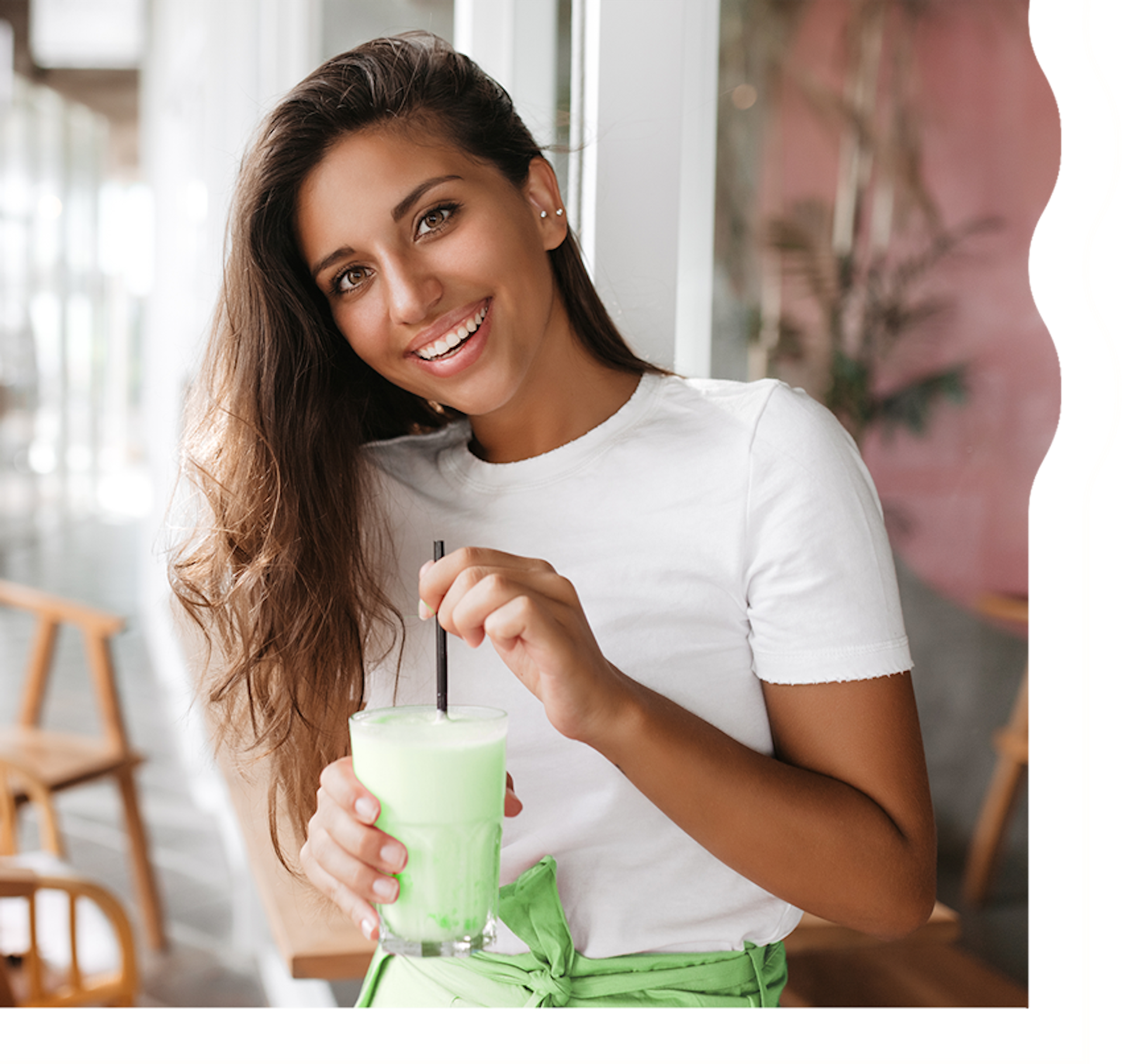 A woman with tan skin and long, dark brown hair is smiling and looking directly at the camera. She is holding a creamy, minty green shake.