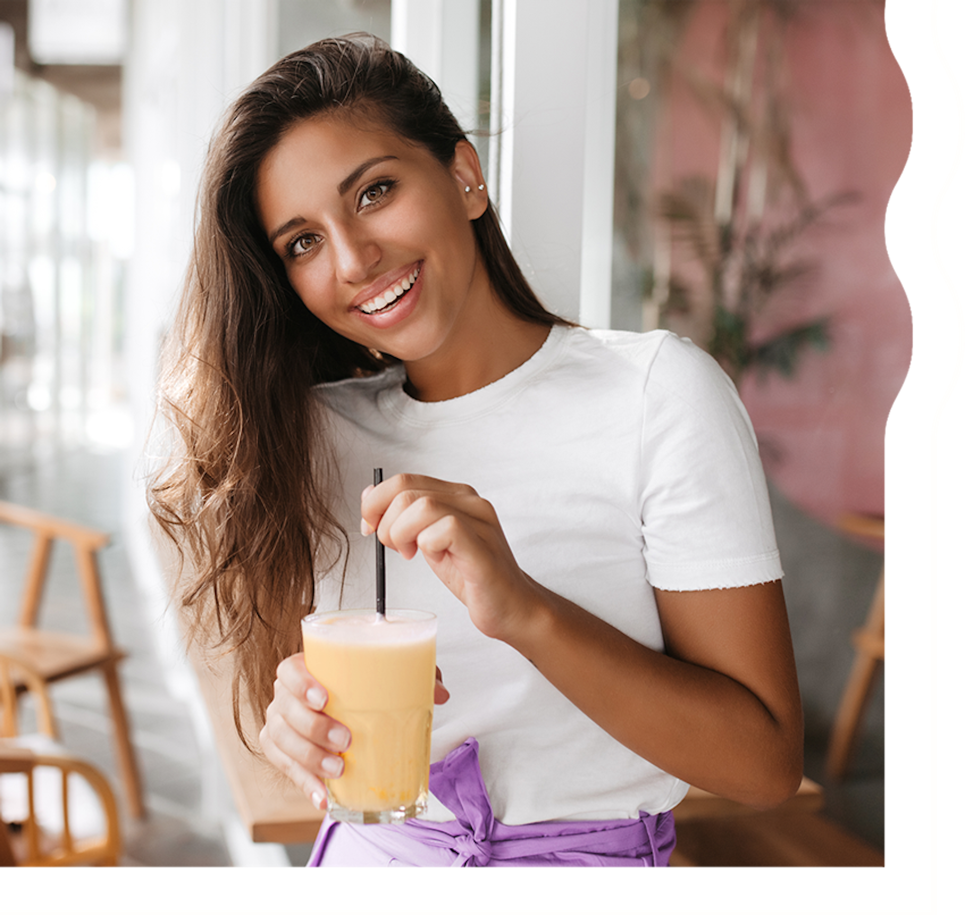 A woman with tan skin and long, dark brown hair is smiling and looking directly at the camera. She is holding a creamy orange shake.