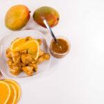 Diced chicken covered in orange sauce on a white plate. Sliced oranges, mangoes, and a jar of orange sauce to the side.